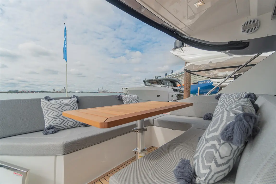 Luxury boat interior featuring a wooden table and grey cushions with tassels, with a marina view and cloudy skies.